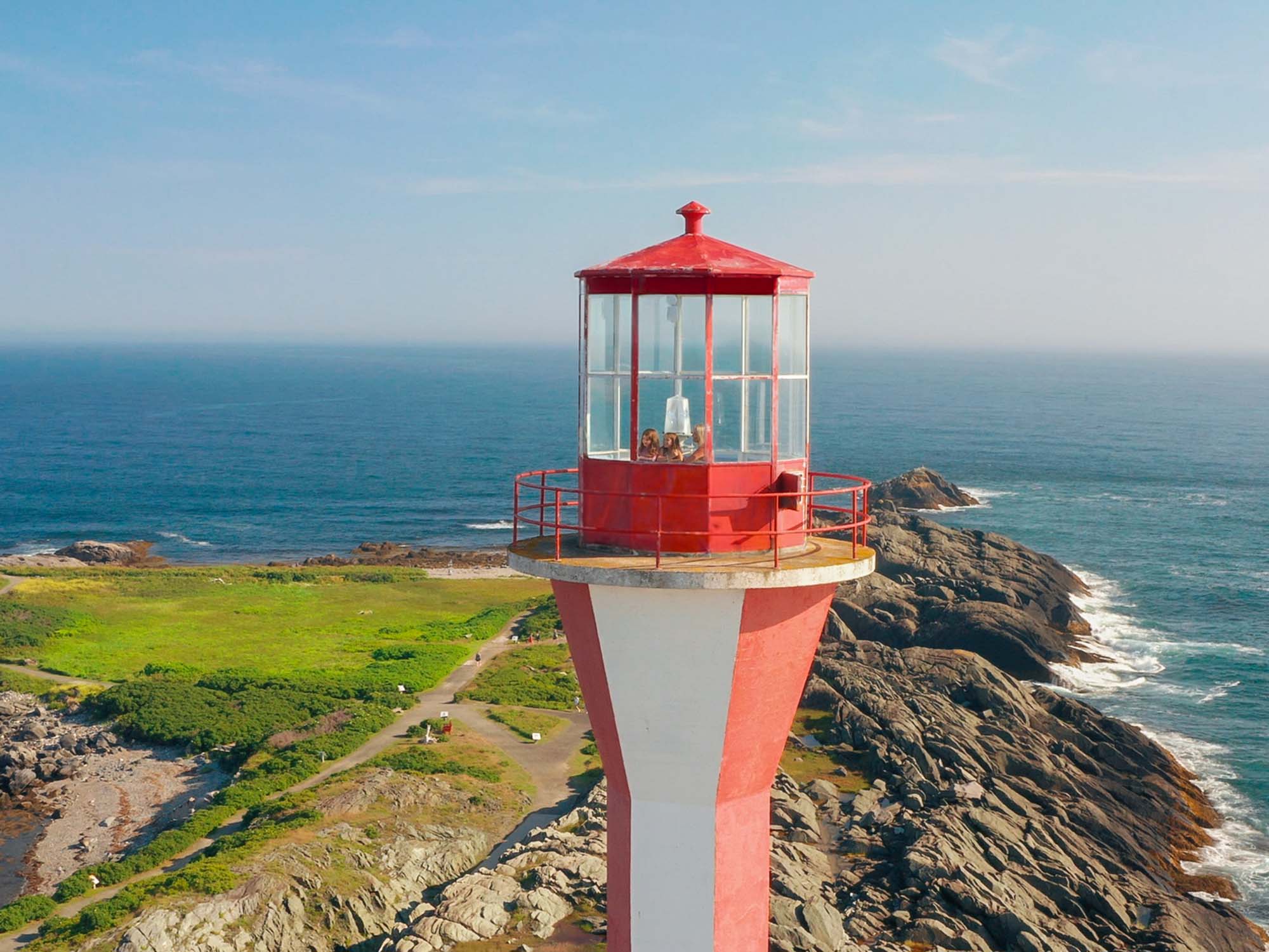 An aerial view of three people inside the top of the Cape Forchu Lighthouse with the rocky coastline, trails and ocean surrounding them on a sunny day near Yarmouth Nova Scotia.